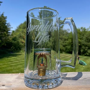 May include: A clear glass beer mug featuring the etched text Sprinkler Life on the side. Inside the mug, a brass fire sprinkler head is mounted in the centre of the base, visible through the transparent glass walls and the sturdy handle.