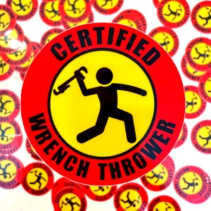 May include: A circular sticker featuring a yellow centre and a red border. The text CERTIFIED WRENCH THROWER is printed in black along the border. In the centre, a black stick figure is depicted in a throwing motion holding a pipe wrench.
