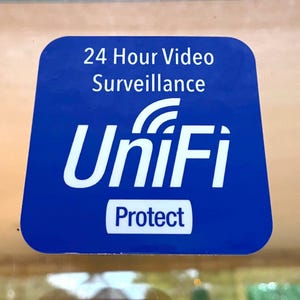 May include: A blue square sticker with white text that reads "24 Hour Video Surveillance" and "UniFi Protect". The sticker also has a white Wi-Fi symbol.