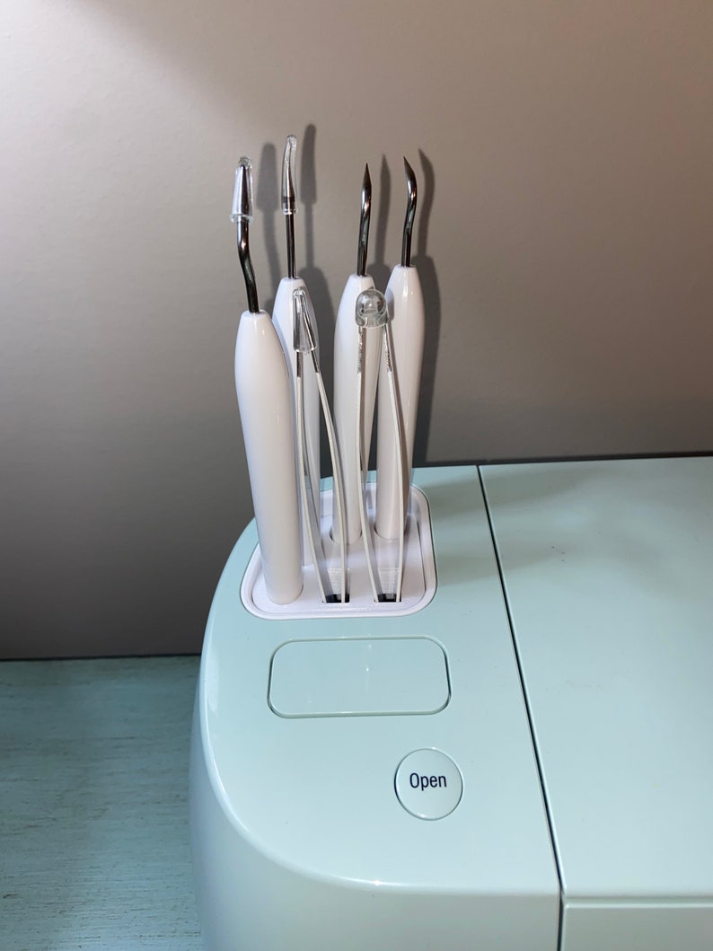 May include: A light blue crafting machine with a white tool holder containing various tools. The machine has a button labeled "Open".