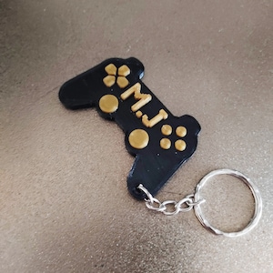 Könnte beinhalten: Schwarzer Plastik-Videospiel-Controller-Schlüsselanhänger mit goldenen Knöpfen und dem Namen "M.J." auf der Seite.