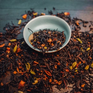 May include: A close-up of a small blue bowl filled with loose leaf black tea, with orange and green pieces of fruit and spices scattered around the bowl on a dark wooden surface.