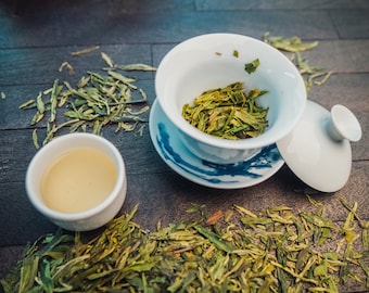 Puede incluir: Una taza de té de cerámica blanca con un diseño azul y blanco, llena de hojas de té verde sueltas. La taza de té está sobre una superficie de madera con hojas de té verde sueltas esparcidas a su alrededor. Una taza de té de cerámica blanca con té amarillo claro también está sobre la superficie.