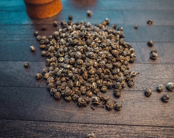 May include: A close-up of a pile of loose leaf green tea pearls on a dark wood surface. The tea pearls are small and round, with a light green colour and a dark brown centre.
