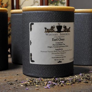 Può includere: Quattro barattoli di tè cilindrici grigi con coperchi in bambù. L'etichetta sul barattolo anteriore recita "Western Immortal Earl Grey" ed elenca ingredienti e origine. Foglie di tè sfuse sparse sulla superficie di legno. Ogni barattolo è alto circa 10 cm.