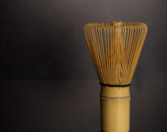 May include: A bamboo matcha whisk, also known as a chasen, with a light brown color. The whisk has many thin tines and a cylindrical handle. A black band secures the tines to the handle. The whisk is used for preparing matcha tea.