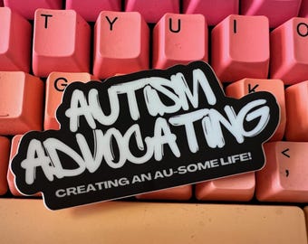 May include: A black and white sticker with the words "AUTISM ADVOCATING" in a bold, stylized font. The sticker also includes the text "CREATING AN AU-SOME LIFE!" The sticker is placed on a keyboard with pink and orange keys.