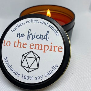 May include: A lit, handmade soy candle in a black tin with a gold interior. The label reads "no friend to the empire" with the scent notes "leather, coffee, and smoke". A geometric shape is also on the label.