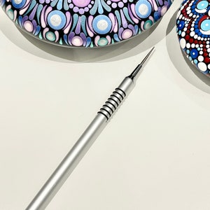 Op de afbeelding: Een zilveren stippenhulpmiddel met een zwarte rubberen grip. Het gereedschap wordt gebruikt om stippen te maken op oppervlakken zoals stenen of canvas.