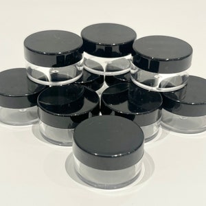 May include: A group of eight small, clear plastic containers with black lids. The containers are stacked on top of each other.