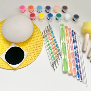 Könnte beinhalten: Ein Bastelset zum Bemalen einer weißen Keramik-Kugel. Das Set enthält 12 kleine Gläser mit Acrylfarbe in verschiedenen Farben, zwei Pinsel, eine gelbe Silikonmatte, einen schwarzen runden Aufkleber und eine Vielzahl von Werkzeugen zum Tupfen und Verwirbeln von Farbe.