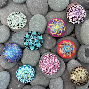 May include: A collection of hand-painted mandala stones in various colors and patterns. The stones feature intricate dot designs in shades of blue, green, pink, yellow, and gold. The stones are arranged on a bed of gray river rocks.