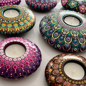 Könnte beinhalten: Ein Set mit sechs handbemalten Teelichthaltern in verschiedenen Farben. Jeder Halter zeigt ein einzigartiges Mandala-Design mit filigranen Details. Die Halter sind aus glattem, rundem Material gefertigt und haben eine kleine Vertiefung in der Mitte, um ein Teelicht aufzunehmen.