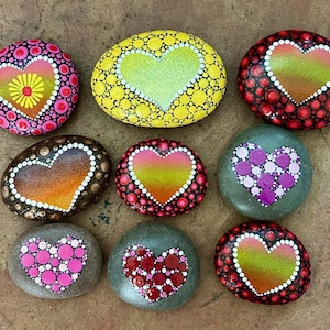 May include: Nine hand-painted decorative stones with heart designs. Each stone features a unique color palette, including pink, yellow, red, and brown, with dot patterns and heart shapes. The stones are arranged on a brown surface.