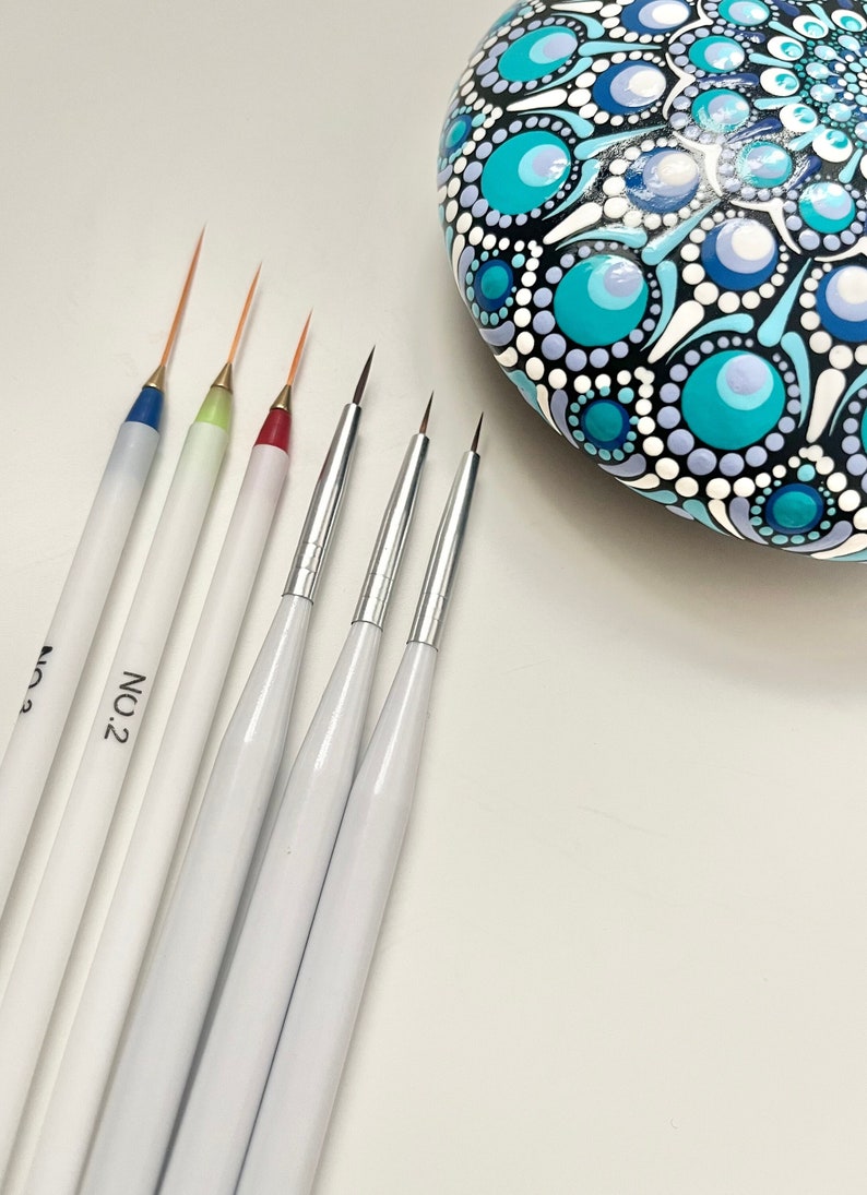 May include: A set of six fine-tipped paint brushes with white handles and gold tips. The brushes are arranged in a row on a white surface. The brush handles are labeled with the number "2". A painted rock with a blue, white, and black mandala design is in the background.