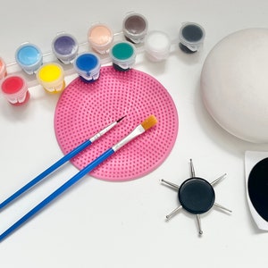 Könnte beinhalten: Ein Bastelset zum Bemalen einer Styroporkugel. Das Set enthält 12 Farbtöpfe in verschiedenen Farben, zwei Pinsel, eine rosa Farbtrocknungsmatte, einen schwarzen Punkt-Aufkleber, eine schwarze Basis mit sechs Metallstiften und eine weiße Styroporkugel.