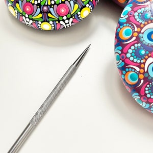 May include: A close-up of a silver dotting tool used for creating dot art on rocks. The tool is lying on a white surface. Three painted rocks with colourful mandala designs are visible in the background.