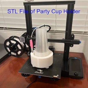 May include: A white 3D printed party cup holder is being printed on a black 3D printer. The printer is on a wooden surface. The text "STL File of Party Cup Holder" is visible at the top of the image.