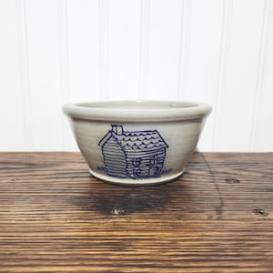 May include: A light gray ceramic bowl with a smooth, rounded rim and a tapered base. The exterior features a dark blue stamped illustration of a small cabin with a chimney, textured roof shingles, and visible window and door outlines.
