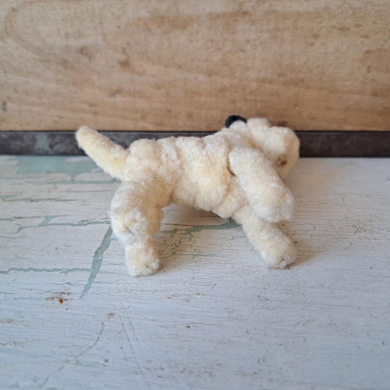 Pu&ograve; includere: Un piccolo giocattolo a forma di cane in peluche, in stile vintage, di colore crema chiaro. Il cane &egrave; in posa giocosa, con la coda sollevata. Il giocattolo ha un naso nero ed &egrave; realizzato in un materiale morbido e strutturato.
