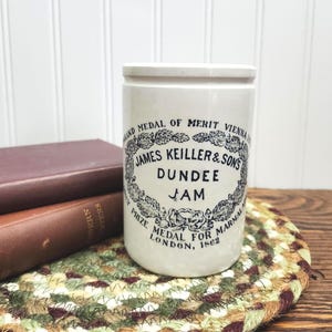 May include: A cylindrical white ceramic jar with black printed text that reads James Keiller & Sons Dundee Jam, Grand Medal of Merit Vienna 1873, and Prize Medal for Marmalade London 1862, surrounded by a decorative wreath of oak leaves.