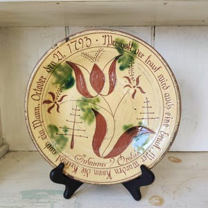 May include: A round ceramic plate with a cream-colored glaze, featuring a hand-painted reddish-brown tulip design, green accents, and stylized tree motifs. The rim is inscribed with German text and the date October 21, 1793, in black script.