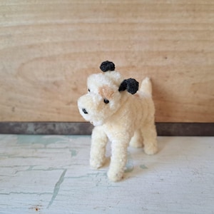 Pu&ograve; includere: Un piccolo giocattolo a forma di cane in peluche, color crema, con orecchie e naso neri. Ha una macchia marrone chiaro sul viso. Il giocattolo &egrave; su una superficie bianca e azzurra.