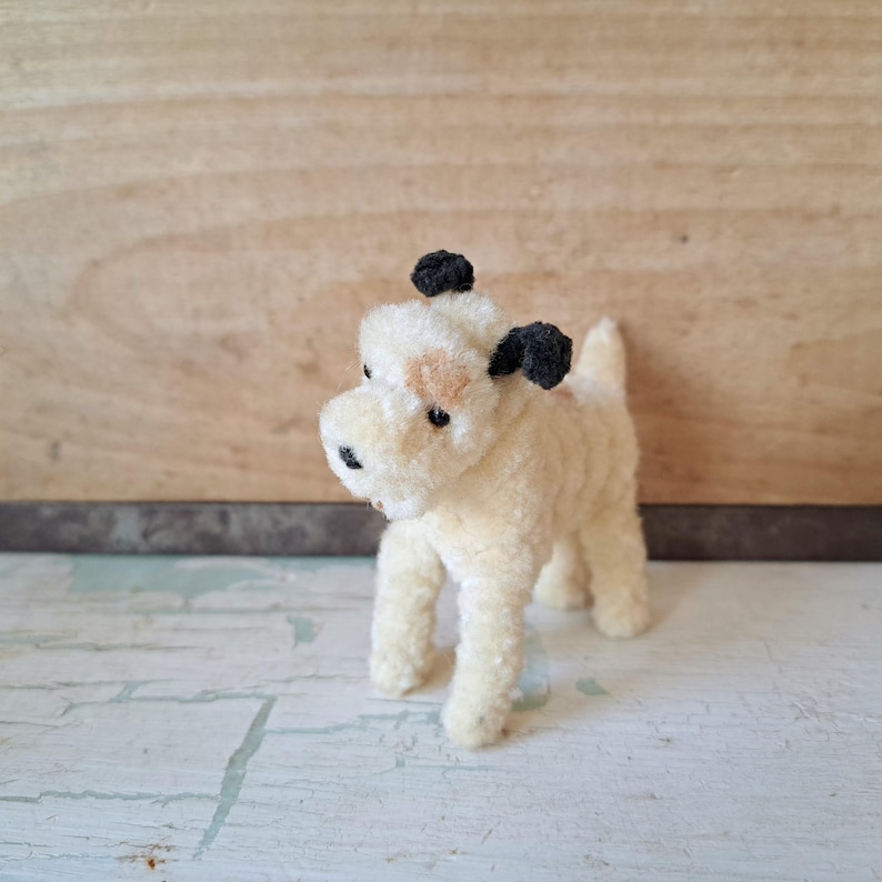 Pu&ograve; includere: Un piccolo giocattolo a forma di cane in peluche, color crema, con orecchie e naso neri. Ha una macchia marrone chiaro sul viso. Il giocattolo &egrave; su una superficie bianca e azzurra.