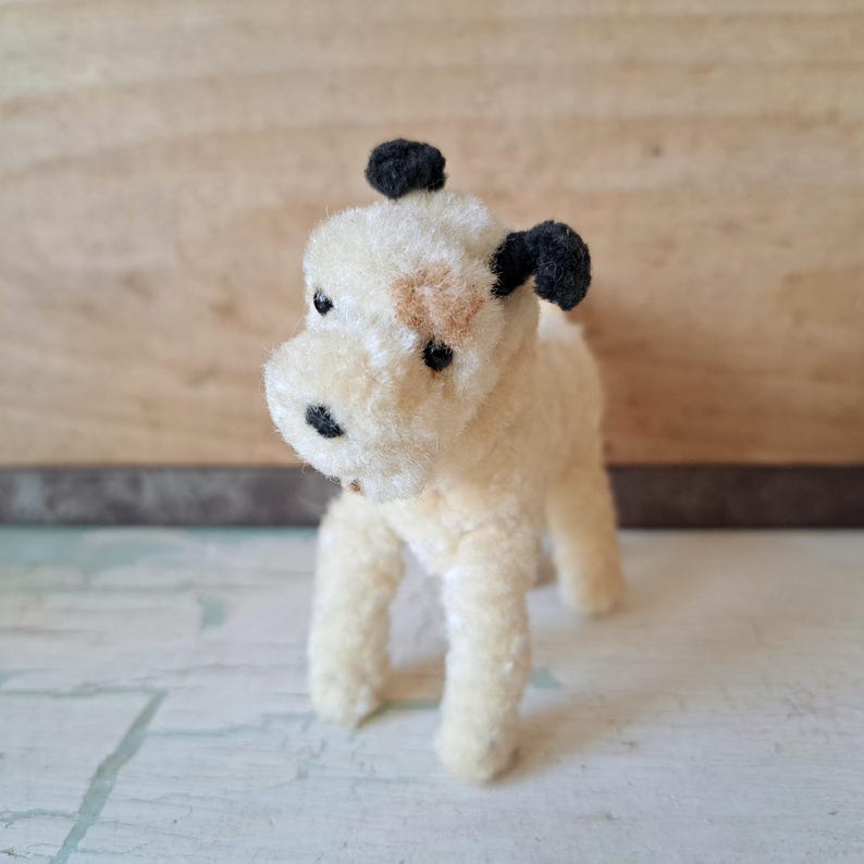 Pu&ograve; includere: Un piccolo giocattolo a forma di cane fatto a mano. Il cane &egrave; color crema con orecchie nere e un naso nero. Ha una macchia marrone sul viso e sta in piedi su quattro zampe. Il giocattolo &egrave; fatto di un materiale morbido e lanuginoso.