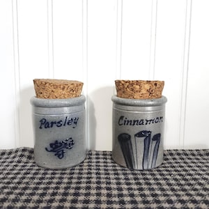 May include: Two cylindrical gray stoneware spice jars with cork stoppers. One jar is labeled Parsley with a blue floral design, and the other is labeled Cinnamon with a blue graphic of three cinnamon sticks. Both feature dark blue lettering.