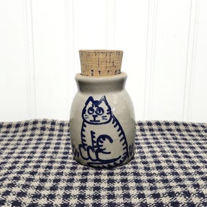 May include: A small, light gray ceramic jar with a cylindrical cork stopper. The jar features a hand-painted, dark blue line drawing of a seated cat on its front surface. The ceramic has a smooth, glossy finish and a rounded, stout body shape.