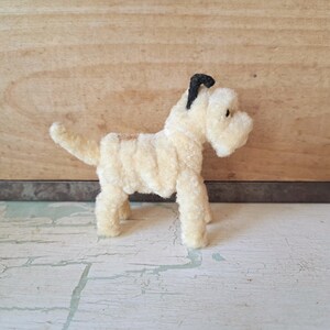 Pu&ograve; includere: Una piccola statuetta di cane color crema realizzata con scovolini sfocati. Il cane presenta un orecchio nero, un piccolo occhio nero e una coda corta e dritta. Il corpo &egrave; costruito con materiale testurizzato avvolto su quattro zampe.