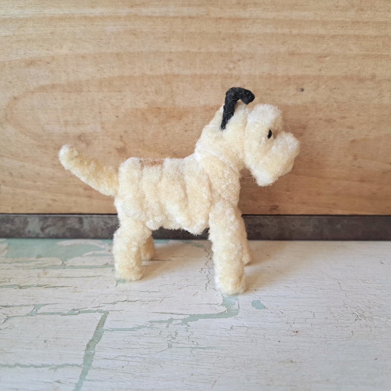 Pu&ograve; includere: Una piccola statuetta di cane color crema realizzata con scovolini sfocati. Il cane presenta un orecchio nero, un piccolo occhio nero e una coda corta e dritta. Il corpo &egrave; costruito con materiale testurizzato avvolto su quattro zampe.