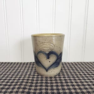 May include: A beige stoneware tumbler with a speckled finish, featuring a blue painted heart design on the front. An oval stamp near the rim reads "PEMAQUID POTTERY" and "BRISTOL, MAINE" with a horizontal line separating the two lines of text.