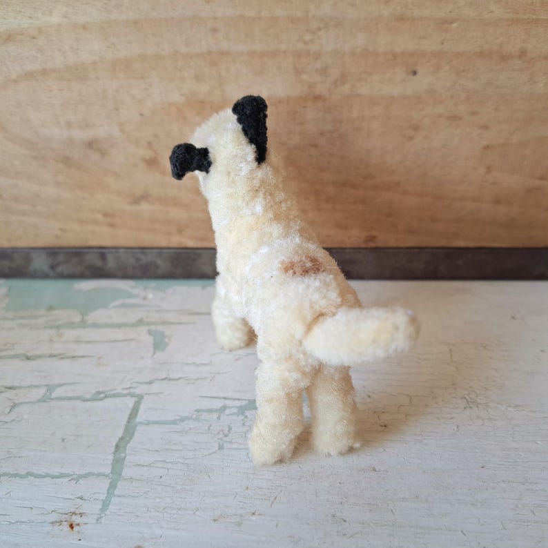 Pu&ograve; includere: Piccola statuetta di cane fatta a mano, con corpo color crema e orecchie e naso neri. Il cane ha una piccola macchia marrone sul fianco e una coda soffice. Il giocattolo &egrave; in piedi sulle zampe posteriori.