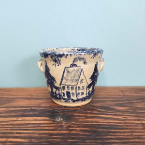 May include: A small, beige ceramic pot featuring a hand-painted blue design of a house surrounded by trees. The vessel has two small side handles and a speckled blue rim. The artwork depicts a simple structure with a chimney and windows.
