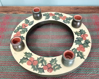 Può includere: Un portacandele circolare in legno con una superficie color crema decorata con foglie di agrifoglio verdi e bacche rosse dipinte a mano. L'oggetto include quattro supporti cilindrici verde scuro con interni color terracotta.