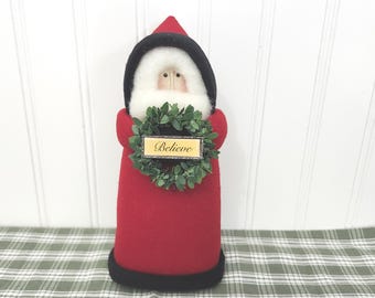 Può includere: Una statuetta di Babbo Natale in tessuto rosso e nero con barba bianca, un cappello a punta e una ghirlanda verde tenuta al centro. La ghirlanda contiene un piccolo cartello rettangolare con la parola Believe in corsivo nero.