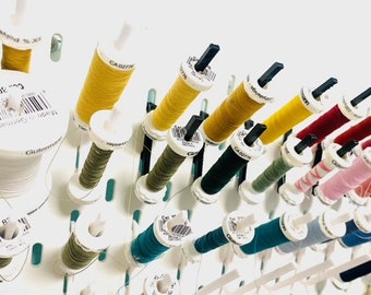 Könnte beinhalten: Nahaufnahme eines weißen Kunststoff-Spulenhalters mit verschiedenen Farben von Garnrollen. Die Spulen sind in Reihen angeordnet und mit Zahlen und Buchstaben beschriftet. Die Garnfarben umfassen Gelb, Grün, Blau, Rot und Pink.