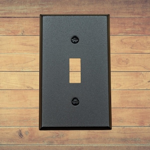 A single black metal toggle switch plate with matching screws installed on a brown wood background.