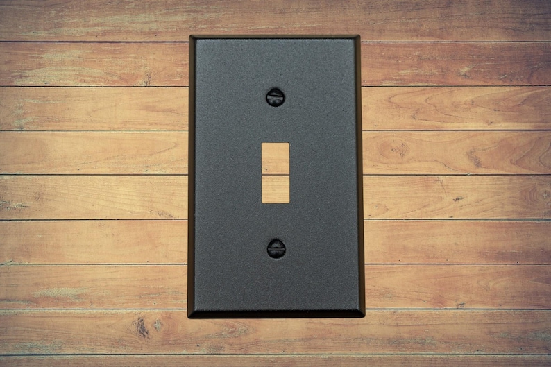 A single black metal toggle switch plate with matching screws installed on a brown wood background.
