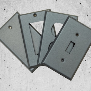 Puede incluir: Cuatro placas de pared eléctricas de metal gris con diferentes configuraciones de interruptores y tomas de corriente. Las placas están dispuestas en forma de abanico.