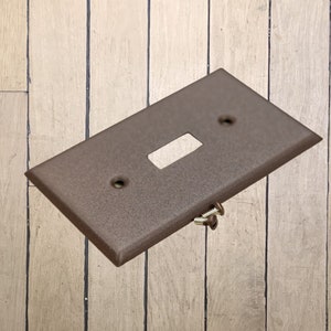 May include: A walnut brown metal single-gang electrical switch plate with two screw holes and a single switch cutout.