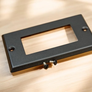 May include: A black metal plate with a rectangular cutout. Two black screws are visible on the bottom edge.
