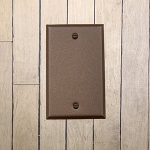 May include: A walnut brown metal electrical outlet cover plate with two screw holes.