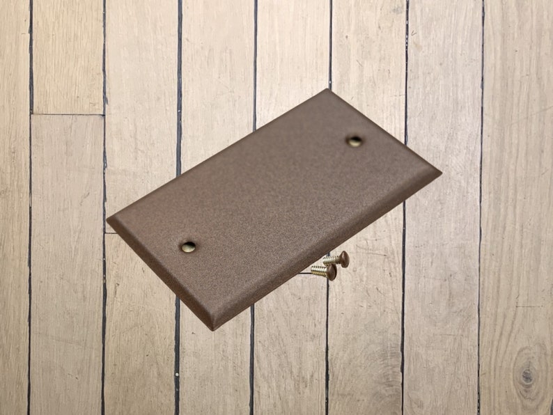 May include: A walnut brown metal wall plate with two screw holes. The plate is lying on a wood surface with two screws next to it.
