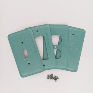 May include: Four teal-colored electrical switch plates with a textured finish. The plates include a single-switch plate, a double-switch plate, and a plate for an electrical outlet. Small screws are included.