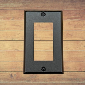 A single black metal rocker switch plate with matching screws installed on a brown wood background.