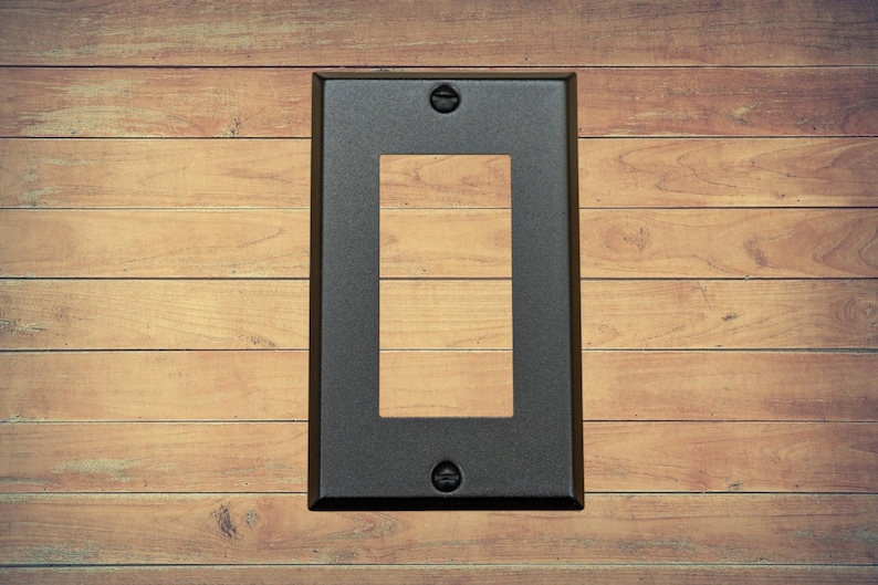 A single black metal rocker switch plate with matching screws installed on a brown wood background.