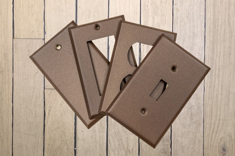 May include: Four walnut brown metal electrical outlet covers with different cutouts. The covers are arranged in a fan shape on a light wood surface.